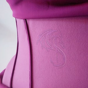May include: A magenta corset featuring a structured design with vertical seams and a smooth fabric finish. A subtle, tonal embroidered dragon motif is visible on the side panel, adding a delicate texture to the vibrant, solid-colored garment.