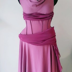 May include: A floor-length mauve dress featuring a cowl neckline, a structured corset-style bodice with vertical seams, and a draped dark magenta sash at the waist. The shoulders are accented with gold-toned circular hardware details.