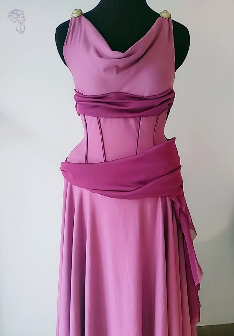 May include: A floor-length mauve dress featuring a cowl neckline, a structured corset-style bodice with vertical seams, and a draped dark magenta sash at the waist. The shoulders are accented with gold-toned circular hardware details.