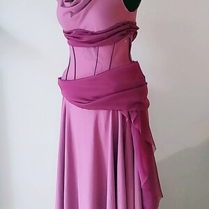 May include: A floor-length mauve dress featuring a draped cowl neckline, a structured bodice with vertical seams, and a darker magenta sash wrapped around the waist. The shoulders are accented with gold-toned circular embellishments.