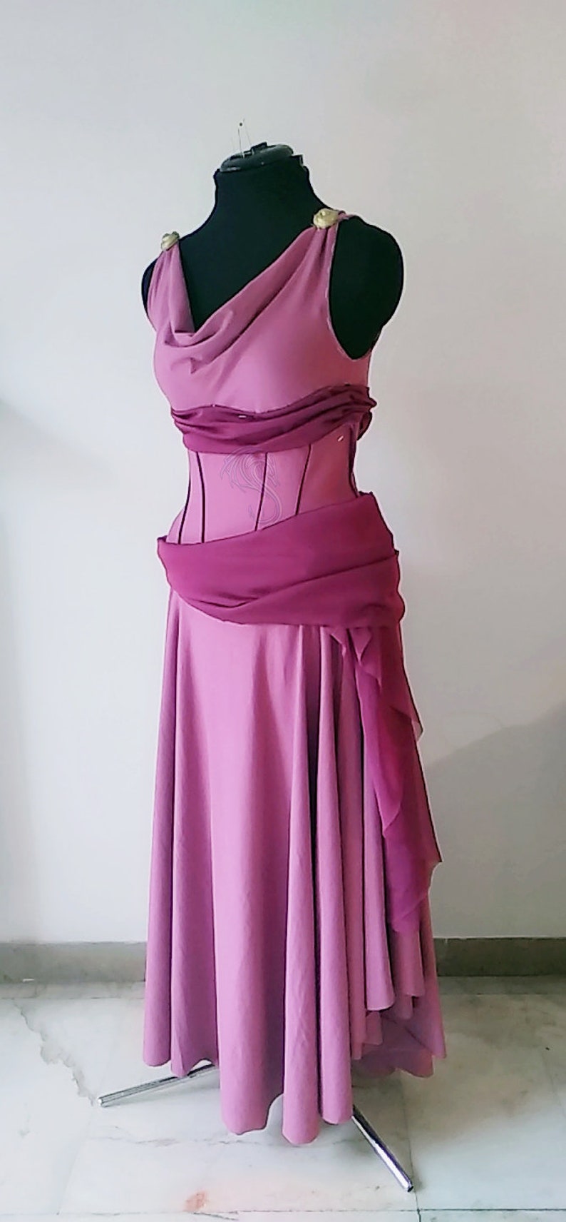 May include: A floor-length mauve dress featuring a draped cowl neckline, a structured bodice with vertical seams, and a darker magenta sash wrapped around the waist. The shoulders are accented with gold-toned circular embellishments.