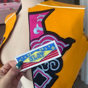 Op de afbeelding: Een oranje vest met witte bies en een kleurrijke patch met de tekst "SLAY". Het vest heeft een zwarte rits en is versierd met roze, blauwe en zwarte borduursels. De rechthoekige patch heeft een geel, rood en blauw ontwerp.