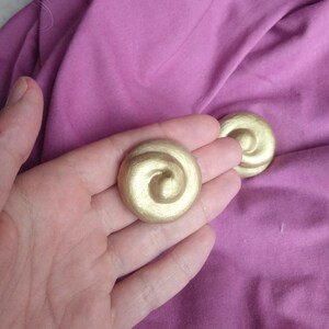 May include: Two circular, metallic gold-colored buttons or decorative accents, each featuring a raised, swirling spiral pattern that starts from the center and radiates outward to the edge, creating a textured, three-dimensional appearance.