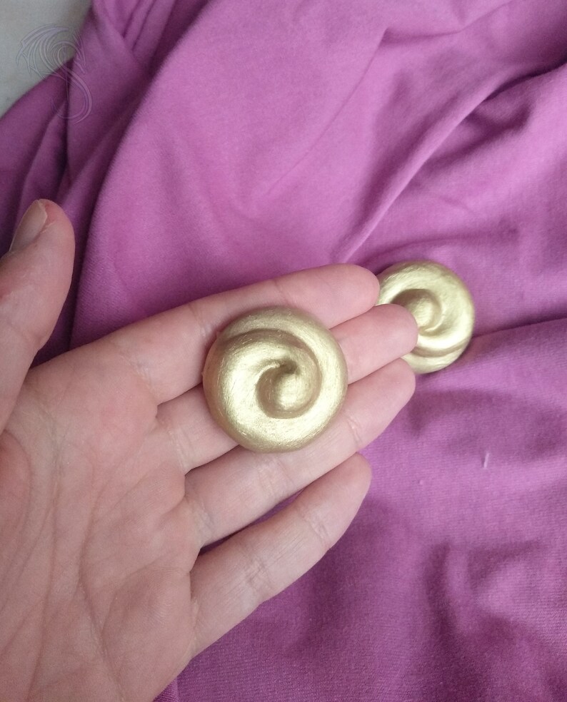 May include: Two circular, metallic gold-colored buttons or decorative accents, each featuring a raised, swirling spiral pattern that starts from the center and radiates outward to the edge, creating a textured, three-dimensional appearance.