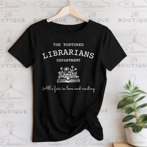 May include: Black t-shirt with white text that reads "The Tortured Librarians Department" and an illustration of an open book with flowers. The text below the book reads "All's fair in love and reading."