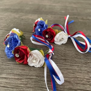 May include: Two red, white, and blue flower crowns with ribbon ties. The crowns are made of fabric flowers with rhinestones in the center of each flower.