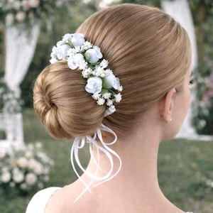 May include: A woman's hairstyle features a low bun adorned with a floral hairpiece. The hairpiece is composed of small, light blue roses and white accent flowers. White ribbons are tied at the base of the bun, adding a delicate touch. The background is blurred.