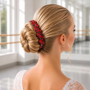 May include: A hair accessory featuring a row of vibrant red, faceted crystals arranged in an elongated, curved shape. The piece is secured into a smooth, blonde hair bun, providing a striking contrast against the light-coloured hair strands.