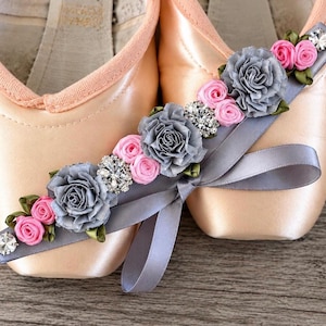 Könnte beinhalten: Ein Paar hellpfirsichfarbene Satin-Spitzenschuhe, verziert mit einem grauen Band, das mit grauen Stoffrosen, kleinen rosa Bandrosetten, grünen Stoffblättern und klaren Kristall-Strasssteinen besetzt ist.