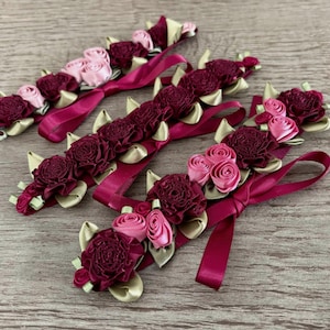 May include: Three decorative bands featuring clusters of fabric roses in shades of deep burgundy and light pink. Each band is accented with small, folded gold-toned fabric leaves and includes a long, smooth burgundy ribbon tie at the end.