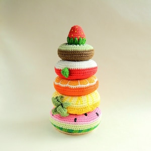 May include: A crocheted fruit-themed stacking toy. The toy features a strawberry, a donut-shaped kiwi, a donut-shaped orange, a donut-shaped lemon, and a donut-shaped watermelon. The colors include red, green, brown, white, orange, yellow, pink, and green.