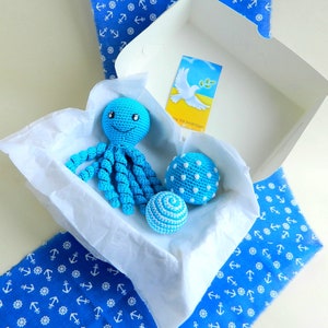 May include: A crocheted blue octopus with a smiling face and curly tentacles rests in a white box with two blue crocheted balls. The box is lined with white tissue paper and sits on a blue fabric with white anchor patterns.