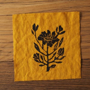 May include: A yellow linen fabric square with a black floral design printed on it. The design features a large flower with five petals and a smaller flower with three petals. The flowers are connected by stems and leaves.