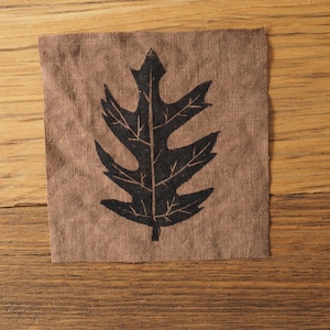 May include: A square, brown fabric patch with a black oak leaf design. The leaf is centered on the patch and has detailed veins. The fabric has a slightly rough texture. The background is a wooden surface.