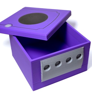 May include: A purple box with a hinged lid, featuring a black circle on top and a silver panel with four circular indentations on the front. The box is open, revealing a matching purple interior.