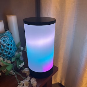 May include: A cylindrical lamp with a black base and a white frosted glass shade. The lamp is lit with a gradient of blue, purple, and white light.