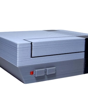 Puede incluir: Una consola de videojuegos retro gris y negra. La consola tiene forma rectangular con una parte superior gris y una base gris más oscura. Cuenta con un panel negro en el lateral y un botón de encendido rojo. Un artículo de juego clásico.
