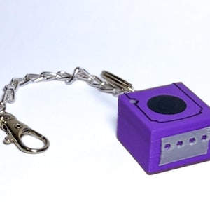 May include: A purple keychain featuring a miniature GameCube design. The cube has a black circle and silver accents. It is attached to a silver chain and a clasp.