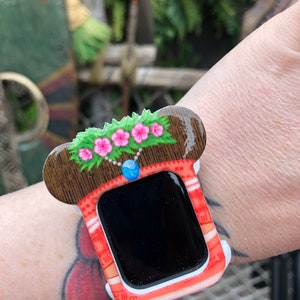 Op de afbeelding: Een wit en rood horlogebandje met een bruin en groen ontwerp. Het horlogebandje heeft een bloemmotief met roze bloemen en een blauwe edelsteen. Het horlogebandje is ontworpen om eruit te zien als een traditionele Hawaiiaanse lei.