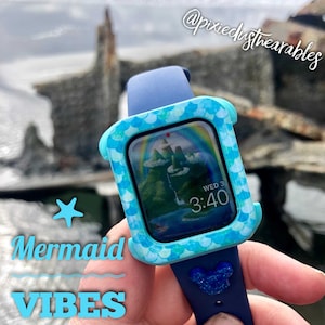 Può includere: Un Apple Watch blu con una cassa dal design a squame di sirena. Il quadrante dell'orologio mostra una scena di montagna colorata con l'ora 3:40 e il giorno WED 3. Il cinturino dell'orologio è blu con un ciondolo di Mickey Mouse glitterato.