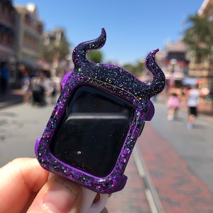 May include: A purple and black glittery watch case with curved horn-like accents. The watch case has a rectangular shape and is held up against a blurred outdoor background.