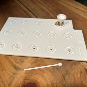 May include: White plastic mold with 16 circular depressions for making earrings. The mold is on a wooden surface. A white plastic tool is also included.