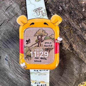 Könnte beinhalten: Eine gelb-rote Smartwatch mit Winnie-the-Pooh-Motiv. Das Zifferblatt zeigt eine Karte und die Uhrzeit 11:29 Uhr mit den Worten „Owls House“ und „100 Aker Wood“. Das Armband hat ein Kartendesign.