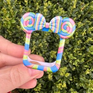 Puede incluir: Un accesorio colorido con temática de caramelos, con una parte superior en forma de lazo y un marco rectangular. El diseño presenta una piruleta en espiral a cada lado del lazo, con un patrón de rayas en tonos rosa, azul, amarillo y verde.