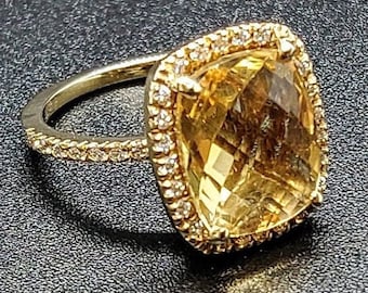 Pode incluir: Um anel de ouro com uma grande gema amarela de forma quadrada, cercada por um halo de pequenas gemas transparentes.