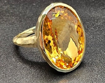 May include: A gold ring with a large, oval-shaped, yellow citrine gemstone. The ring has a hammered texture and a bezel setting.