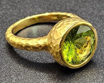 May include: A gold ring with a hammered band and a large, round, green peridot gemstone.