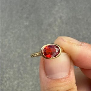 May include: A gold ring featuring an oval-cut, deep red gemstone set in a bezel setting. The ring band has a textured, hammered finish, providing a contrast to the smooth, polished surface of the central red stone and the surrounding gold metal.