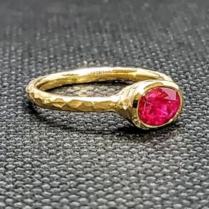May include: A gold ring with a hammered texture band and a bezel-set, round, bright red gemstone. The ring is set against a dark, textured background.