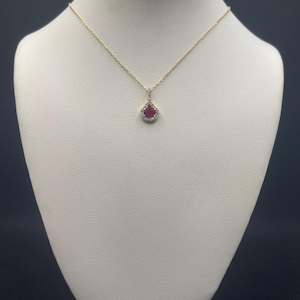 May include: A gold-colored necklace with a teardrop-shaped pendant. The pendant features a red gemstone surrounded by small, clear stones. The necklace is displayed on a white bust against a dark background.