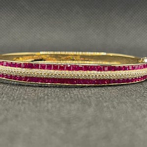 Puede incluir: Una pulsera dorada con una fila de piedras preciosas rojas de corte cuadrado y una fila de piedras más pequeñas y transparentes. La pulsera tiene un cierre y descansa sobre una superficie gris oscuro.