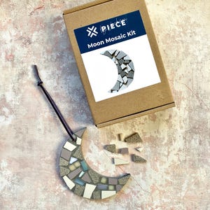 May include: A brown cardboard box with the text "PIECE by piece" and "Moon Mosaic Kit" on the front. The box contains a crescent moon shaped mosaic kit. The mosaic is made of small, irregular pieces of ceramic tile in shades of grey, white, and beige. The mosaic is partially assembled, with some pieces missing. The box is open and the mosaic is sitting on a light brown surface.