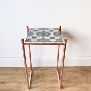 May include: A small side table with a copper-coloured metal frame and a patterned top. The tabletop features a repeating geometric design in shades of grey and white. The table's legs are angled, and the overall design is modern and minimalist.