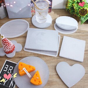 May include: A set of 9 gray concrete coasters in various shapes, including round, square, heart, and oval. The coasters are made of a textured material and have a smooth, polished finish. The coasters are perfect for protecting surfaces from scratches and spills.