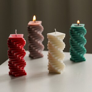 Può includere: Quattro candele decorative di diversi colori, ciascuna realizzata con forme quadrate impilate. Le candele sono disposte in fila su una superficie bianca. Le candele sono rosse, bianche, verdi e viola.