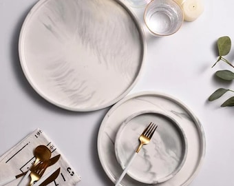 Può includere: Tre piatti rotondi in ceramica bianca con un motivo a marmo grigio. I piatti sono annidati uno dentro l'altro, con il piatto più grande in basso e il piatto più piccolo in alto. I piatti sono su una superficie bianca.