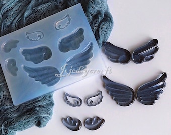 Puede incluir: Un molde de silicona con varias formas de alas, junto con varias alas de resina terminadas. El molde es azul claro y las alas son de un gris oscuro translúcido. La imagen está sobre una superficie blanca con una tela turquesa en el fondo.