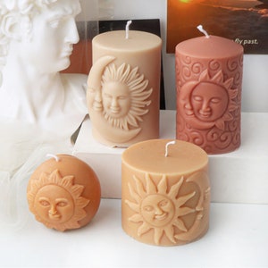 Op de afbeelding: Vier decoratieve kaarsen in beige en koraaltinten. De kaarsen zijn cilindrisch en bolvormig, met ontwerpen van zon- en maan gezichten. De kaarsen hebben witte lonten en zijn waarschijnlijk gemaakt van was.
