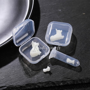 Może przedstawiać: Cztery przezroczyste plastikowe pojemniki z białymi figurkami psów w środku. Pojemniki są kwadratowe i mają pokrywki. Jeden pojemnik jest otwarty i figurka jest widoczna. Pozostałe trzy pojemniki są zamknięte. Jest też przezroczysta plastikowa rurka z białą figurką psa w środku.