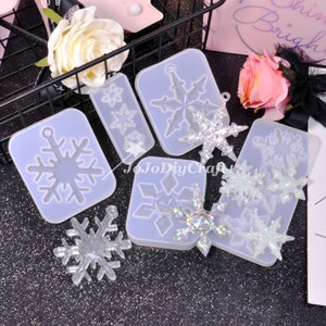 May include: A set of four clear silicone moulds for making snowflake ornaments. The moulds are on a white surface with a few finished snowflake ornaments made with glitter.