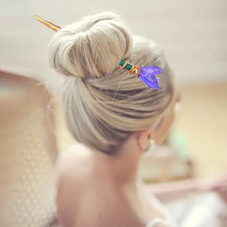 Puede incluir: Una varita para el cabello dorada con un dise&ntilde;o de flor morada y verde. La varita se utiliza para sujetar un mo&ntilde;o en su lugar.
