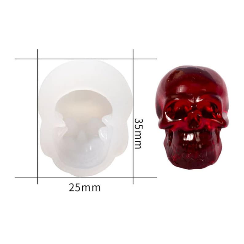 K&ouml;nnte beinhalten: Eine wei&szlig;e Silikonform in Form eines Sch&auml;dels, 6,35 cm breit und 8,89 cm hoch. Neben der Form befindet sich ein roter Glassch&auml;del.