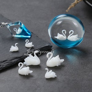 Könnte beinhalten: Ein Set aus weißen Schwanenfiguren, einige sind klein und andere größer. Es gibt auch eine Glaskugel mit zwei weißen Schwänen darin und einen blauen und weißen Kristallanhänger mit einem Schwan darin.