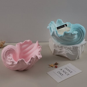 May include: Two decorative seashell-shaped candle holders. One is light blue and has a white candle inside. The other is pink and empty. Both are on a white surface. The text "HAVE A NICE DAY." is on a white card in the foreground.