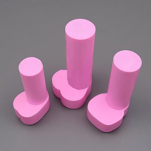 May include: Three pink silicone molds, each featuring a cylindrical top section attached to a rounded, clover-shaped base. The molds vary in height, with one taller central piece and two shorter pieces positioned on either side, all in a solid pink.