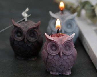 May include: Three owl-shaped candles, one lit, in shades of purple, gray, and brown. The candles are made of wax and have detailed features, including eyes, beaks, and feathers.